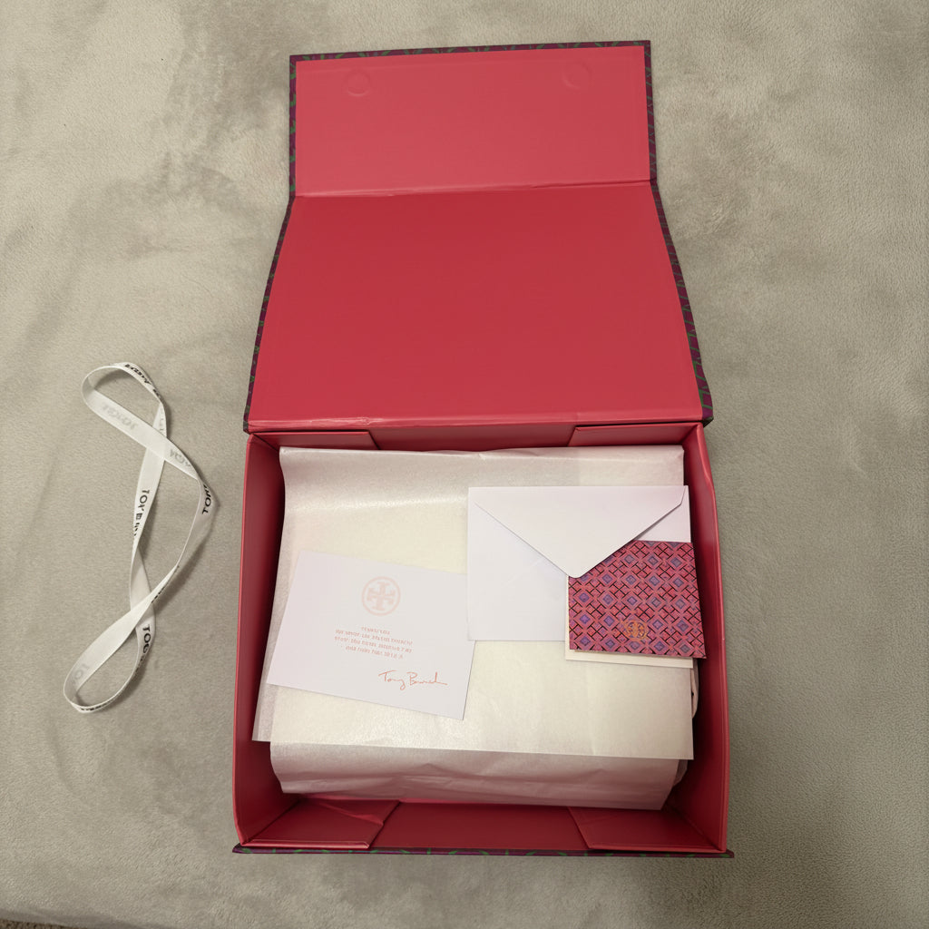 An open red luxury gift box containing white tissue paper, a white card with a circular logo and 'Tory Burch' script, a white envelope, and a small patterned red and gold card on a gray carpet.